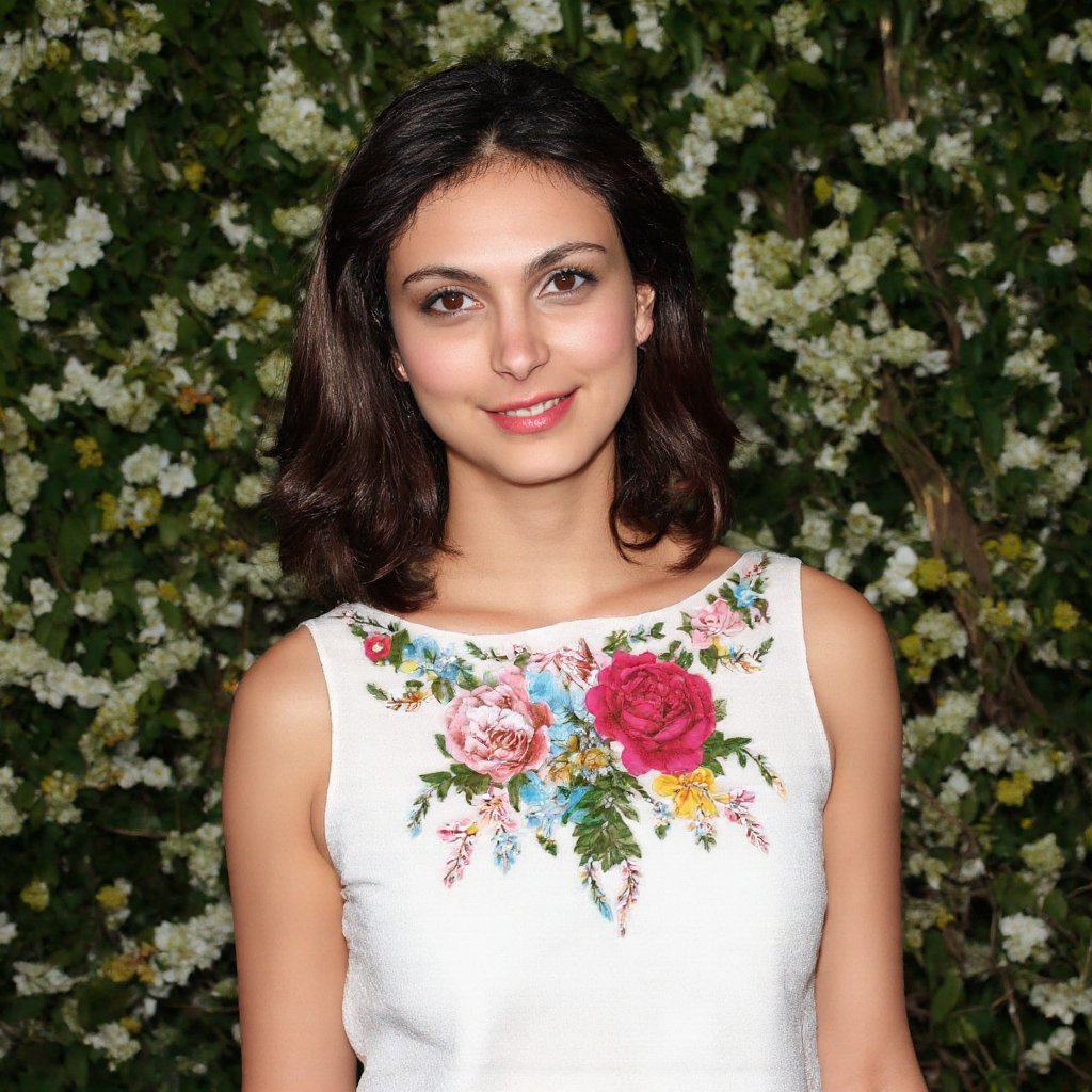 morena_baccarin, A woman is standing in front of a bush. The bush has small white flowers on it. The woman is wearing a white dress with colorful flowers on the front of it. 