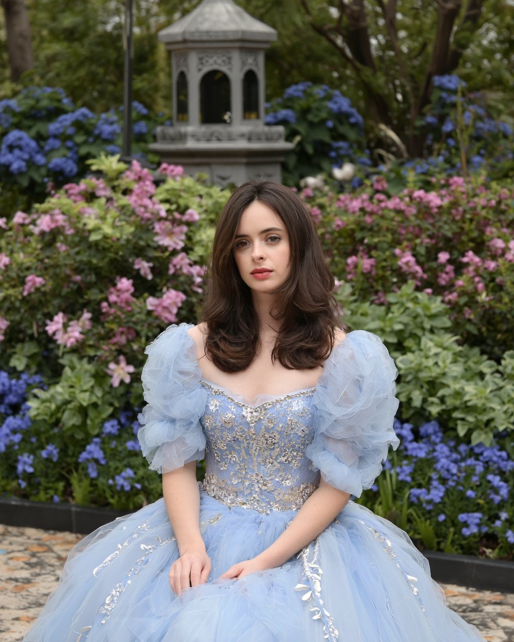 A portrait of krysten_ritter seated in a lush garden. She wears a voluminous, light blue gown with delicate floral patterns and a beaded waistband. Her long, wavy brown hair flows down her back, and she gazes directly at the camera with a contemplative expression. The garden is filled with vibrant flowers in shades of pink, blue, and green, and a geometric lantern rests in the background. The ground is tiled with a mix of geometric and floral patterns. The overall color palette is soft and harmonious, dominated by the blue of her gown and the greens of the plants.