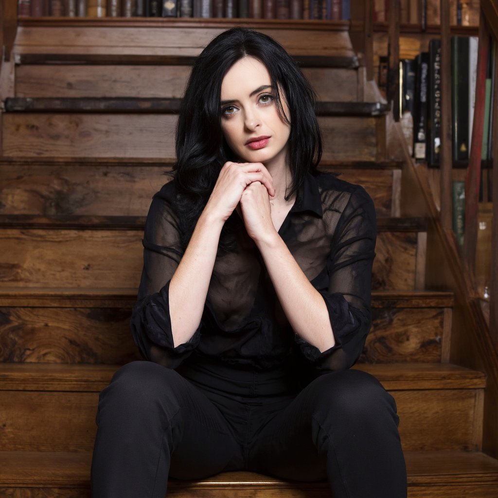 A portrait of krysten_ritter seated on a wooden staircase. She wears a black, sheer blouse and dark trousers. Her hands are clasped in front of her face, and she has a contemplative expression. The background shows a bookshelf filled with books, indicating a library or study. The color palette consists primarily of dark browns, blacks, and muted reds, creating a somber and introspective atmosphere.