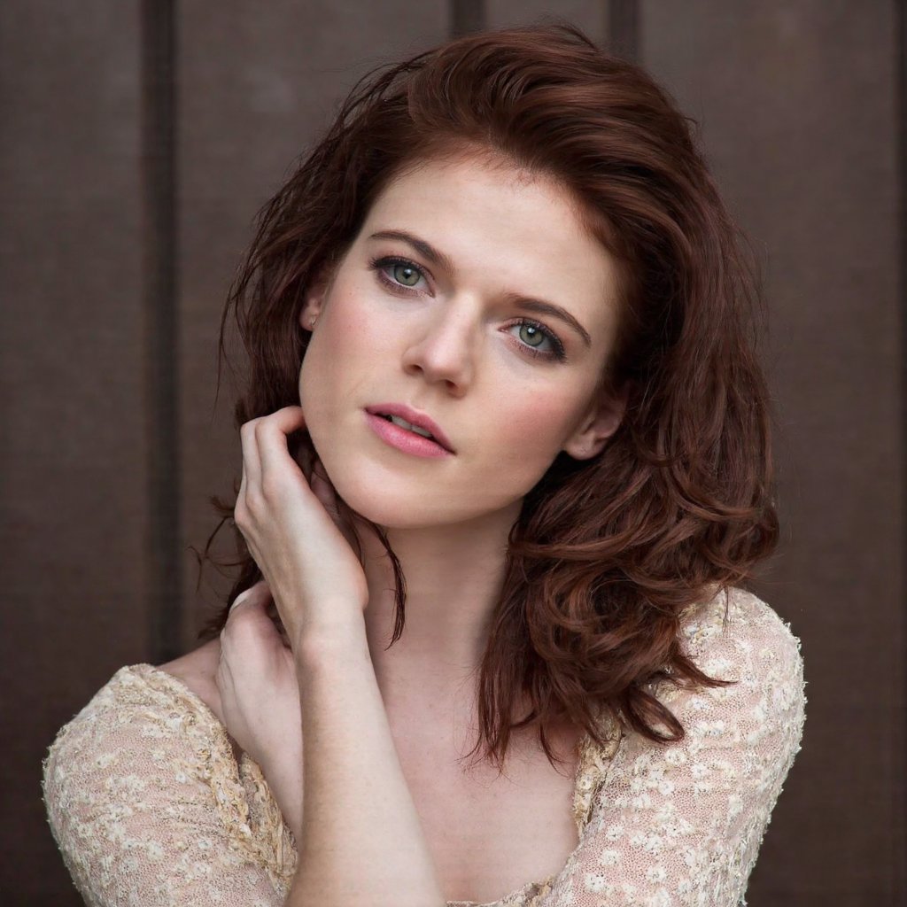 A close-up portrait of rose_leslie with a contemplative expression. She has dark, wavy hair and striking green eyes. Her fair skin is accentuated by subtle makeup, including a light pink lip color. She wears a delicate, off-white dress with intricate lace detailing. The woman's pose is relaxed, with her hand gently touching her neck. The background is a muted brown, likely a wooden door or wall, indicating an indoor setting. The image style is candid and natural, capturing a moment of introspection.