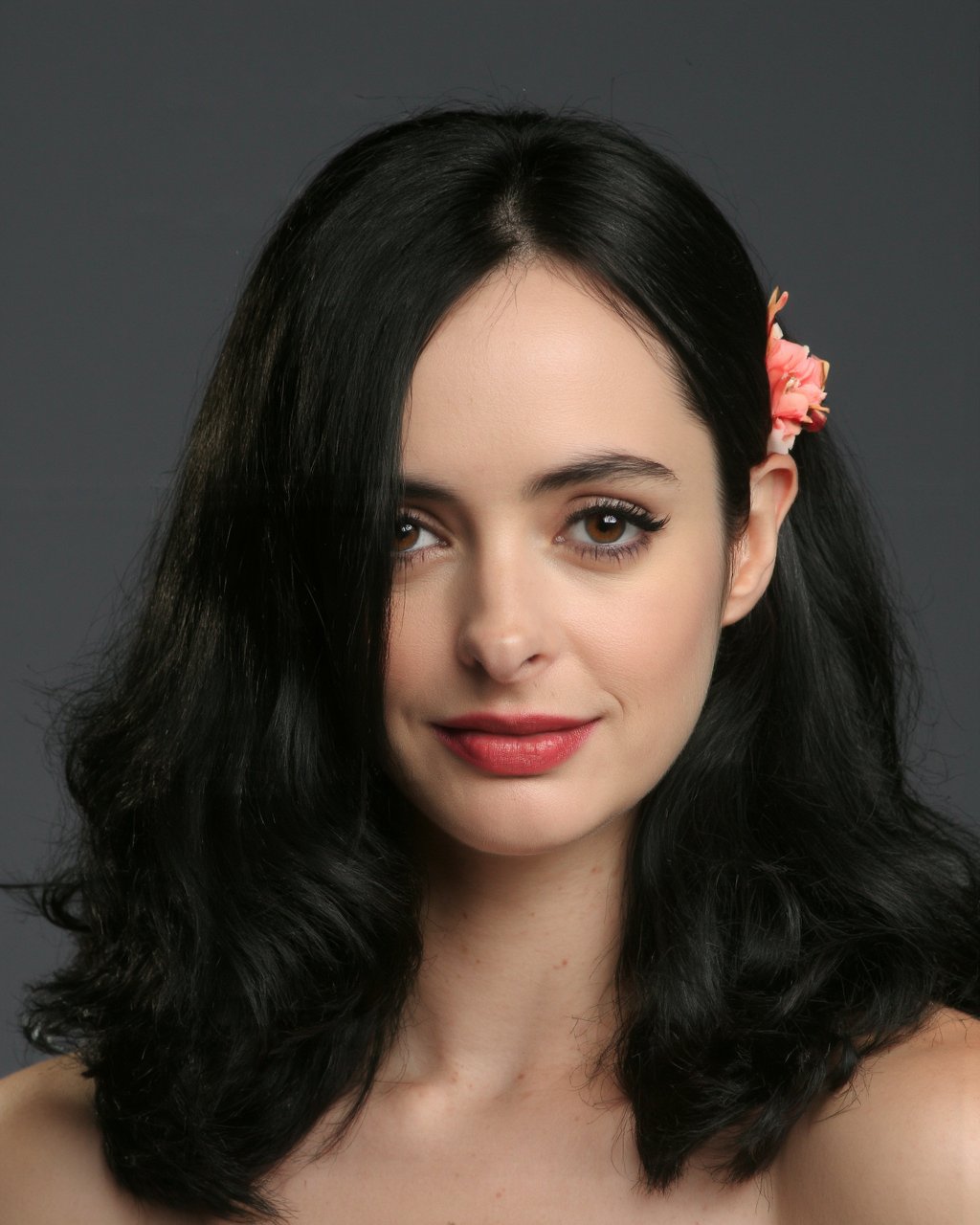 A portrait of krysten_ritter with a serene expression. She has long, dark hair styled in a half-up, half-down manner, secured with a delicate, peach-colored flower. Her makeup is subtle, emphasizing her eyes and lips. The background is a dark, muted gray, contrasting with the woman's pale complexion and the flower's soft pink hue. The image style is elegant and sophisticated, emphasizing the subject's natural beauty and elegance.