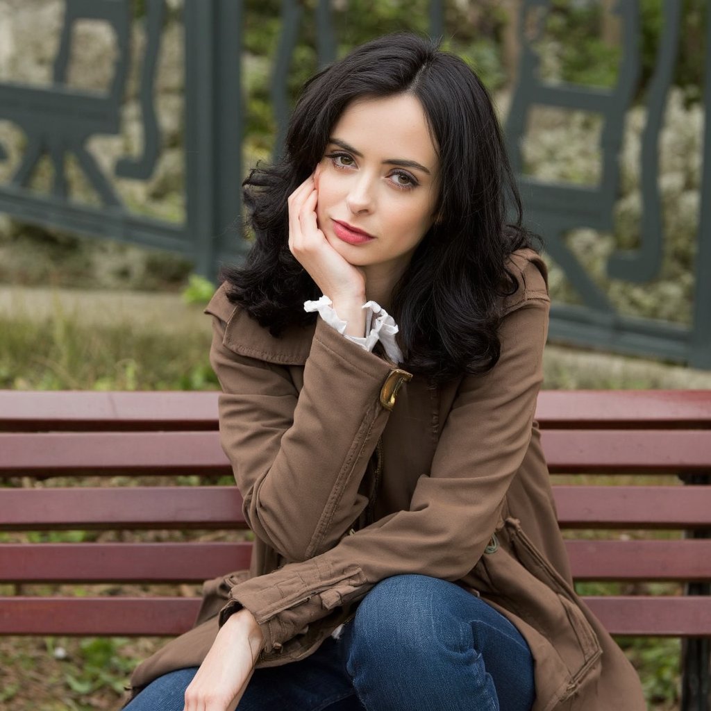 A portrait of krysten_ritter seated on a wooden bench. She has long, wavy brown hair and wears a brown coat over a white blouse. Her pose is contemplative, with one hand resting on her chin and the other on her knee. The bench is a deep red, contrasting with the muted tones of the surroundings. The background features a wrought iron railing with a greek key pattern. The image conveys a serene and introspective mood, with the woman's contemplative expression suggesting introspection.