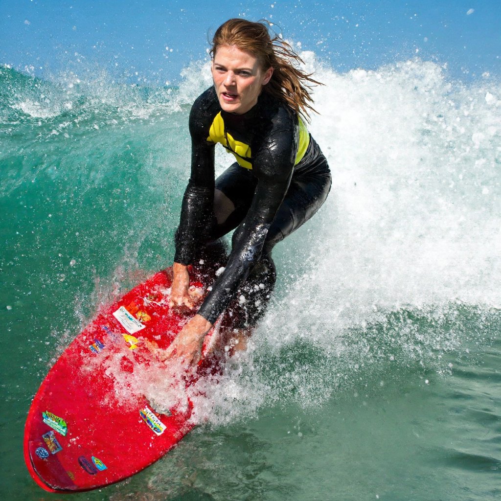 A dynamic close-up action shot of rose_leslie riding a wave. She is wearing a black and yellow wetsuit, is captured mid-ride on a vibrant red surfboard adorned with colorful stickers. Her right arm is extended forward, and her left arm is bent at the elbow. The wave behind the surfer is a brilliant turquoise, with white foam splashing around it. The clear blue sky indicates a sunny day. The image captures the thrill and energy of surfing.