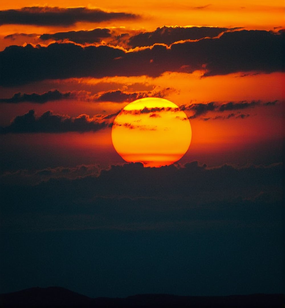 Majestic sunset with the sun engulfed in clouds,vivid orange and black color palette,silhouette of the horizon,atmospheric mood,warm tones,centered composition,prominent sun with visible solar texture,possibly captured with a telephoto lens,minimalist aesthetic,inspired by natural landscape photography,high quality,