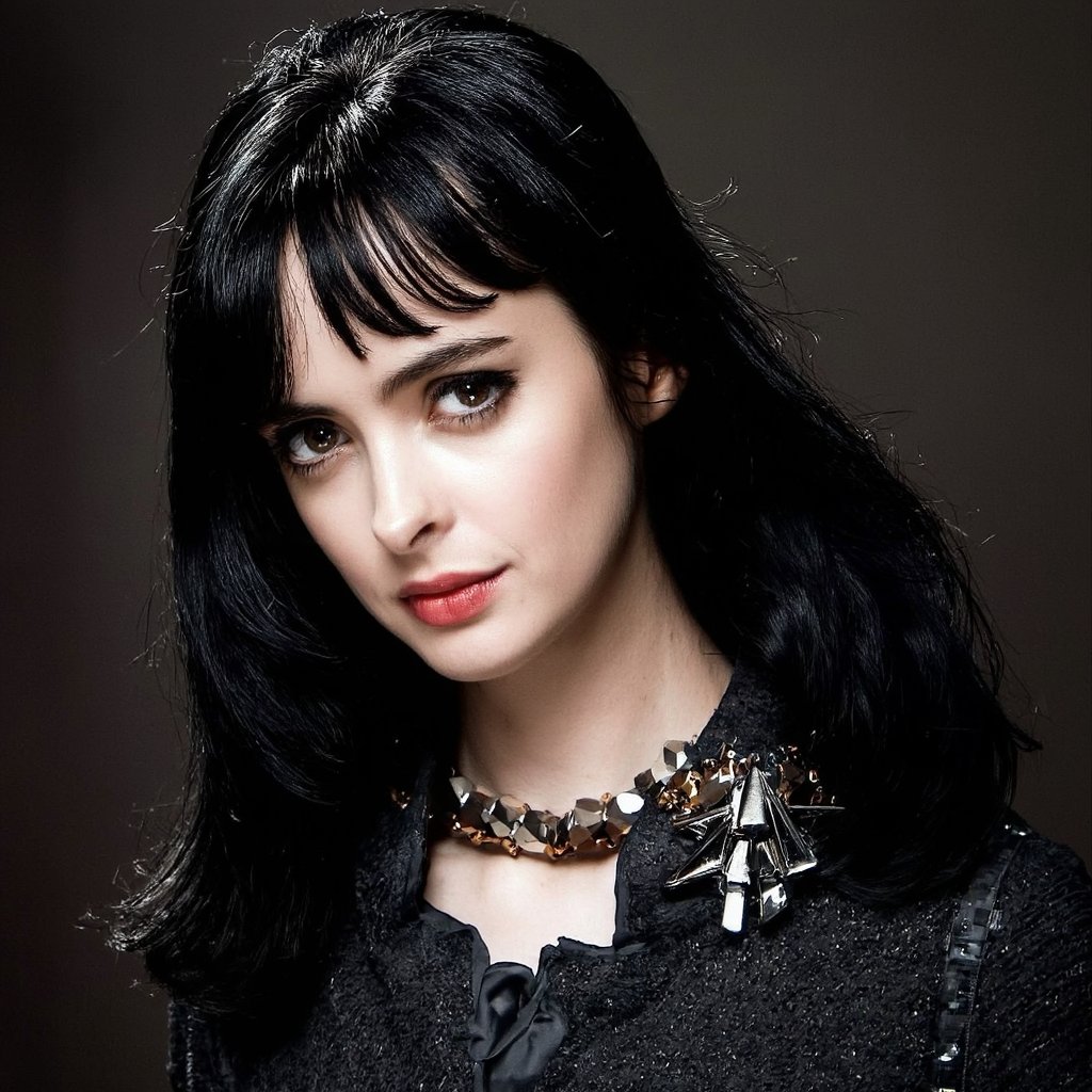 A portrait of krysten_ritter with striking eyes, set against a dark, moody background. Her hair flows freely, creating a sense of movement and energy. She wears a black, textured top and a chunky, metallic necklace with a square-shaped pendant. Her makeup is bold and dramatic, emphasizing her eyes and lips. The image style is contemporary, emphasizing fashion and glamour.