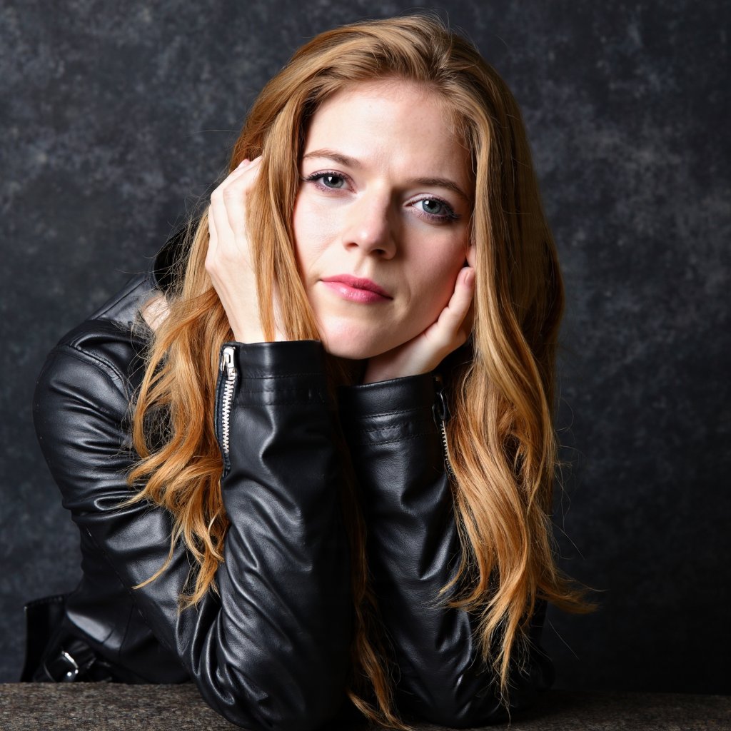 A portrait of rose_leslie with long, wavy blonde hair. She wears a black leather jacket with a zipper. Her left hand rests on her face, and her right hand gently touches her forehead. Her makeup is subtle, with a hint of pink lipstick. The background is a textured, dark-colored surface, possibly a stone or marble wall. The image conveys a serene and contemplative mood.