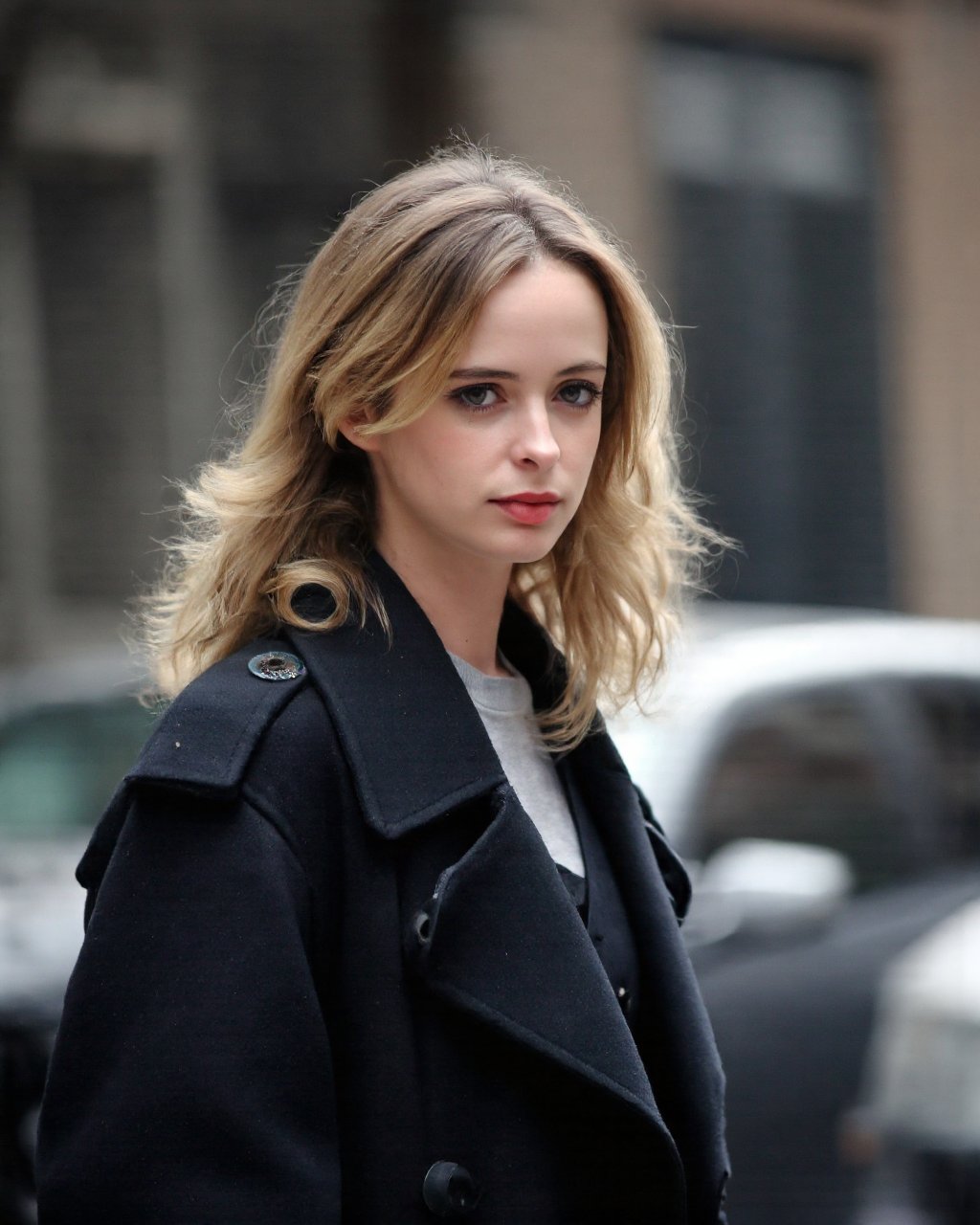 A portrait of krysten_ritter with long, wavy blonde hair, captured in a side profile. She wears a dark, oversized coat. The setting is an urban street with a building visible in the background. Soft lighting casts a gentle glow on her face, highlighting her features and the texture of her hair. The color palette is muted, with the blonde hair contrasting against the darker clothing. The image conveys a serene and contemplative mood.