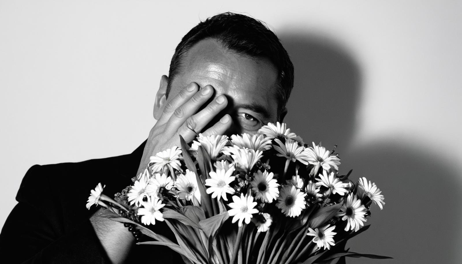 A black and white photograph of a man's face partially obscured by a bouquet of flowers. The man is holding the flowers with both hands in front of his face.  Only his left eye and part of his nose are visible. The man is illuminated from the left, and is wearing a beaded bracelet on his left wrist.