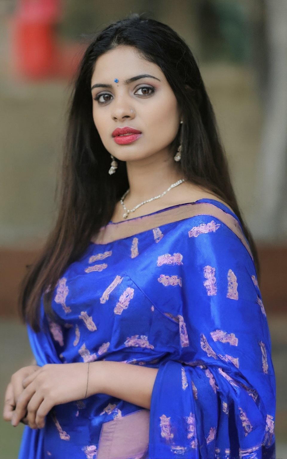 The image contains a person wearing a blue sari with a pink pattern. The individual has long, dark hair and is adorned with a gold necklace featuring a central blue stone. Additionally, there is a ring on the person's right hand. The background appears to be an outdoor setting with greenery.

Her long black hair flows naturally, framing her face, with soft pink lips adding a touch of color to her look. A delicate necklace adorns her neck, and matching earrings provide a subtle accent to her ensemble. The background is complex yet designed to draw attention to her, possibly featuring a shopping environment that suggests a day out. The setting is realistic, with careful attention to the textures of her clothing, the shine of her hair, and the soft glow of her skin, creating a vibrant, relatable scene that emphasizes modern casual fashion.,Mallu beauty ,Mallu ,Realistic photography ,Indian 