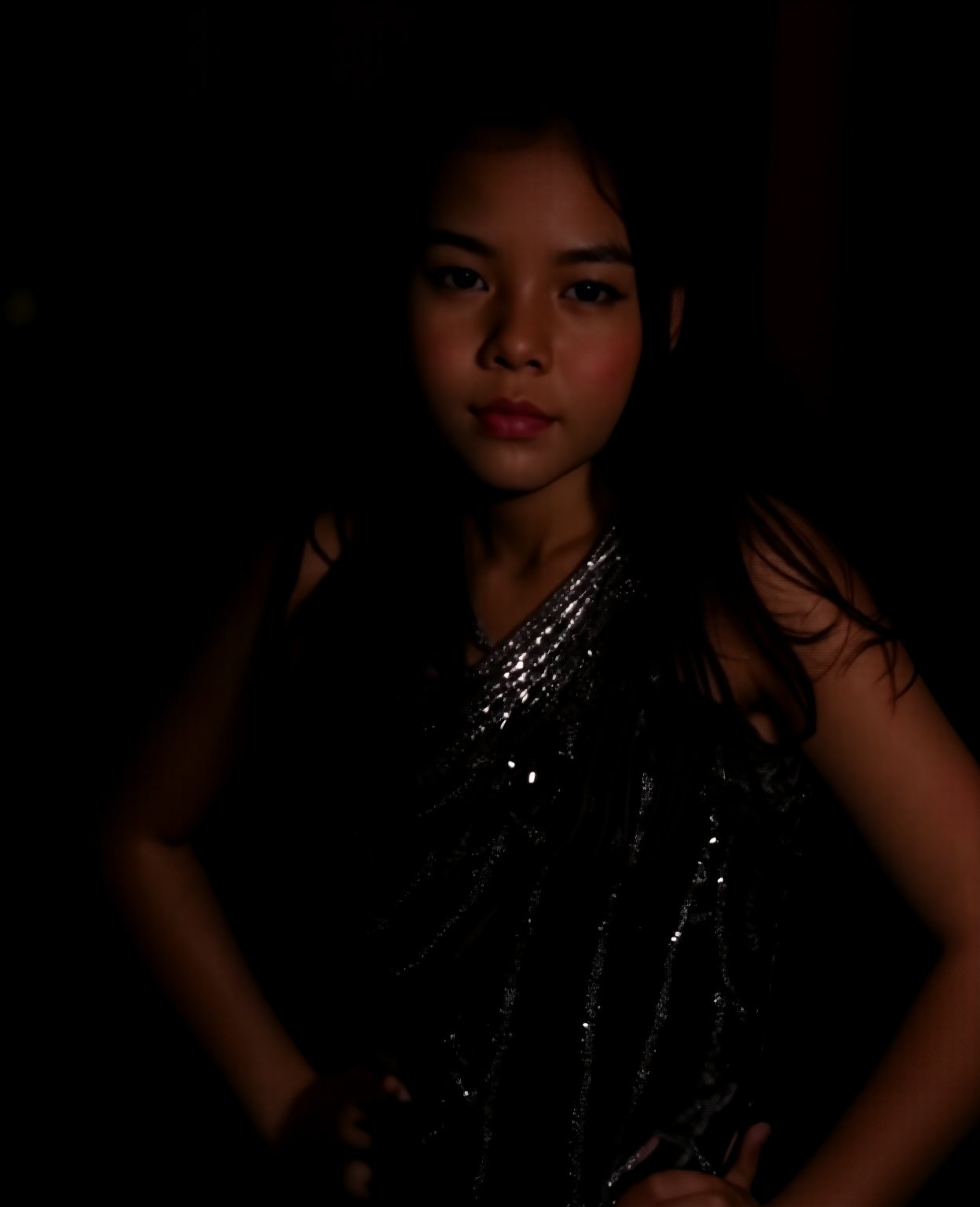 A close-up shot of a young girl standing in a dimly lit room, wearing a sleek black dress that hugs her curves. The dress is adorned with intricate silver sequins that catch the faint light, giving off a subtle sparkle. Her long dark hair cascades down her back like a waterfall, framing her heart-shaped face. She stands confidently, one hand resting on her hip, as if savoring the moment