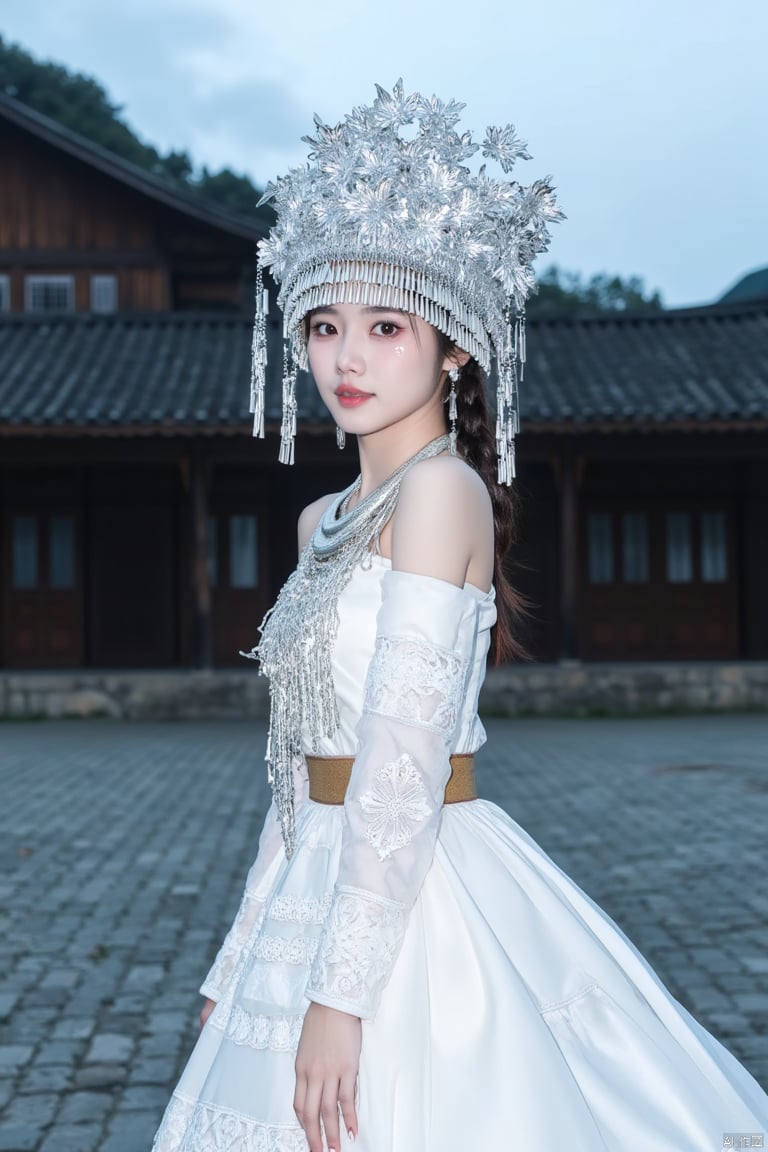 A young woman wearing a silver Miao ethnic headdress,a woman dressed in a white dress and a silver crown adorned with flowers, stands on a cobblestone street. The woman's dress is adorned with white embroidery, adorned with a gold belt. Her hair is pulled back in a ponytail, adding a pop of color to her face. The backdrop, a rustic wooden house, is visible in the distance. The sky is a muted blue, with a few wispy clouds, adding depth to the scene.