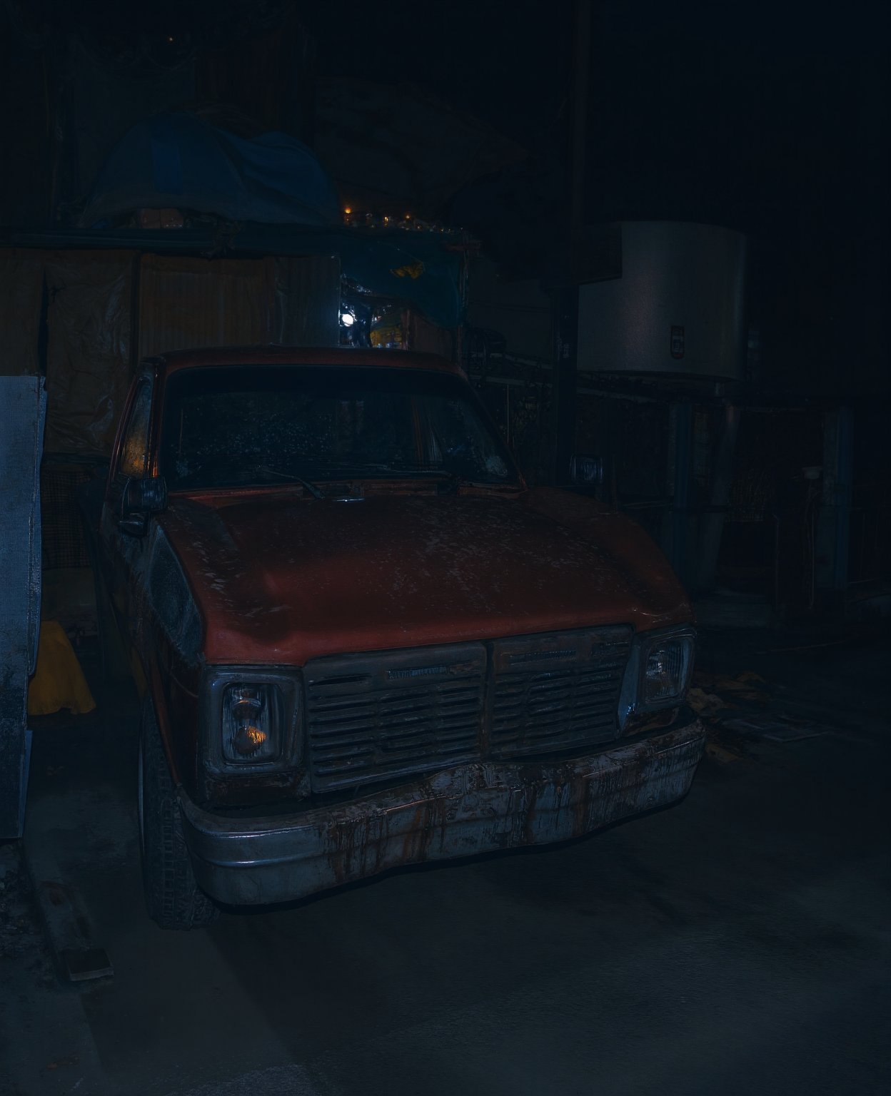 A truck parked on a deserted street at night, its headlights casting an eerie glow. The darkness of the surrounding buildings and the absence of any nearby streetlights emphasize the isolation of this late-night setting. The truck's metal body reflects the faint city lights, giving it a metallic sheen.