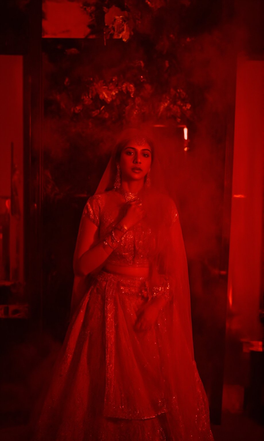 In a dimly lit, smoke-filled room, Madonna Sebastian poses majestically against a lavish, crimson-hued backdrop adorned with gleaming gold accents and subtle neon highlights. Her stunning red lehenga shimmers with intricate embroidery, paired with opulent gold jewelry that catches the eye. The cinematic lighting casts a warm, moody glow, evoking the essence of the iconic Matrix franchise. Madonna Sebastian exudes confidence and glamour, her regal presence commanding attention in this striking, neo-noir inspired setting.