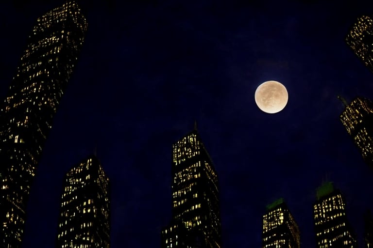 night, dark blue sky with blurry white clouds, full moon, black rectangular skyscrapers with small windows in which white light is visible, metal superstructures are visible at the tops of skyscrapers, brown cubes and a few gray cubes are visible at the base of skyscrapers, the earth is brown mud or clay,