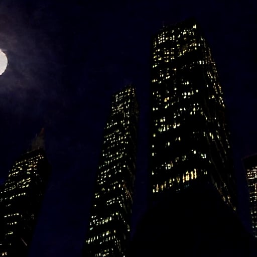 night, dark blue sky with blurry white clouds, full moon, black rectangular skyscrapers with small windows in which white light is visible, metal superstructures are visible at the tops of skyscrapers, brown cubes and a few gray cubes are visible at the base of skyscrapers, the earth is brown mud or clay,