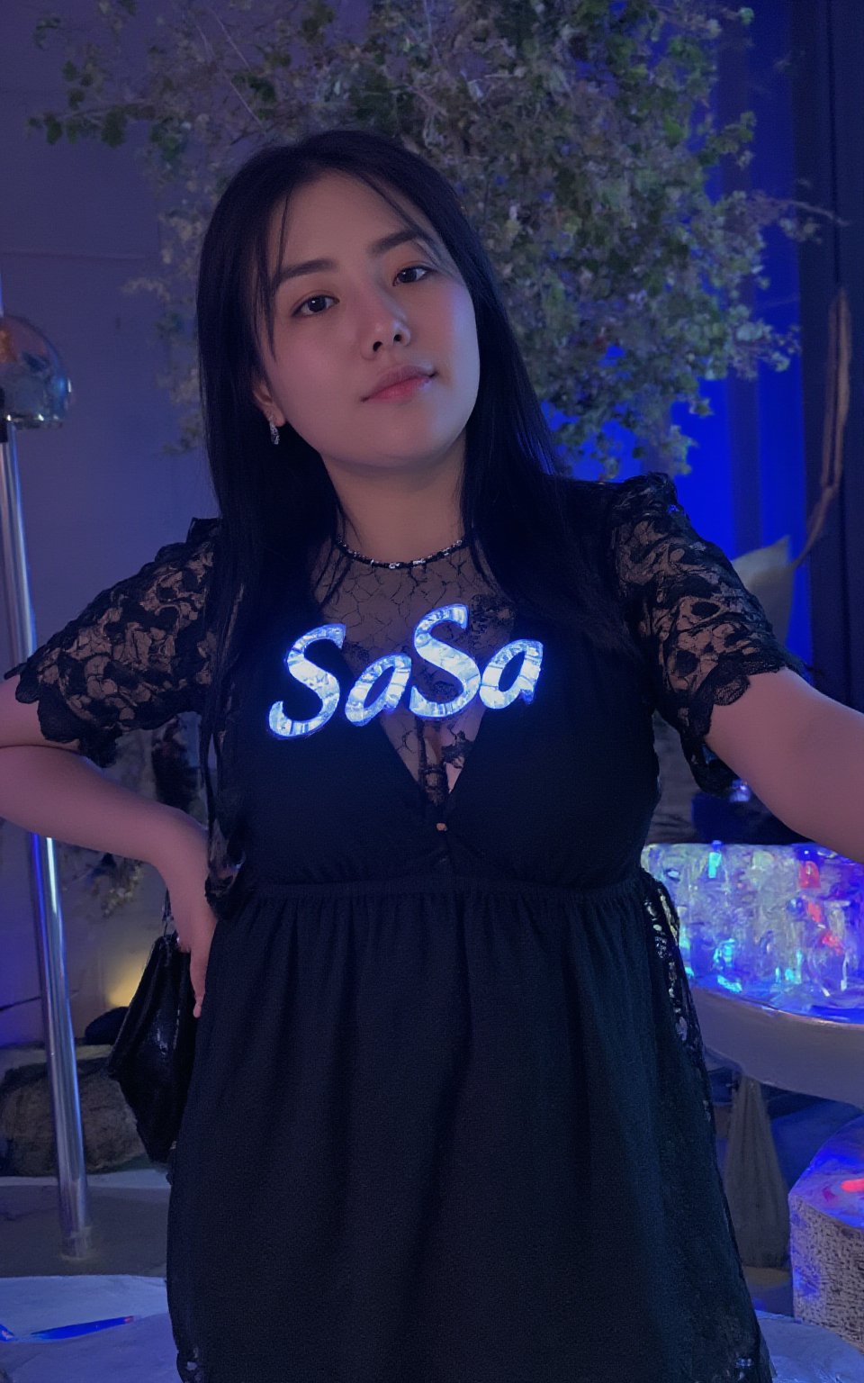 a hubgwoman with long dark hair, wearing a black lace dress, adorned with a necklace and earrings. The woman's chest is adorned with blue letters that spell out "SaSa" in a glowing light-up font. The backdrop is a vibrant blue, with a few colorful lights in the distance.