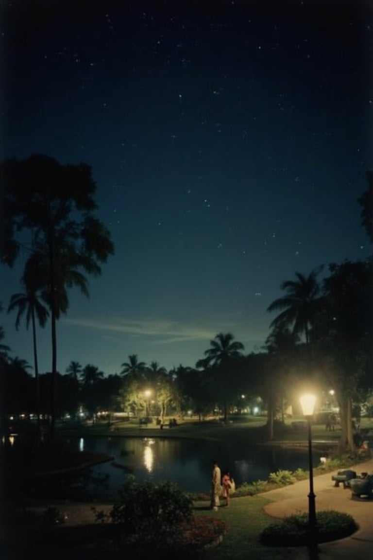 A 1990s nighttime view of Lake Garden in Malaysia, with the scene bathed in soft, ambient lighting. The lake is calm, reflecting the dim glow of nearby street lamps and the starry sky. The garden is serene, with trees and bushes casting long shadows. A few visitors stroll along the paths, their figures barely visible in the low light. The atmosphere is peaceful and tranquil, capturing the essence of a 1990s Malaysian night at Lake Garden.