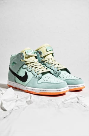 The image features a pair of Nike SB Dunk High Pro sneakers. The shoes are displayed against a crumpled white background. The design of the sneakers is a mix of pastel colors, including a pale yellow toe cap, a soft pink midsole, and a light grey upper section. The shoes have a black Nike swoosh logo on the side, and the laces are in a pale green and light blue color. The sole of the shoes is a solid orange color.