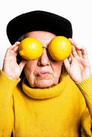 This image depicts an elderly individual wearing a black cap and a yellow sweater. The person is holding two lemons up to their eyes, playfully positioning them as if they were spectacles. The lemons are held over the eyes, one over each eye, creating a humorous effect. The individual's face is partially visible, showing a puckered mouth, which adds to the whimsical nature of the scene. The background is plain and white, which draws attention to the subject and the lemons. The overall mood of the image is light-hearted and fun, with a touch of playfulness.