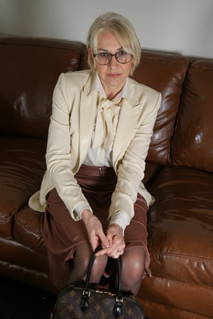 A close-up shot captures the elegance of an older woman seated on a worn brown leather couch, surrounded by subtle hints of moisture. Her cream-colored wool blazer, adorned with a white bow tie and shirt, clings to her body, while the high-waisted pencil skirt falls in soft folds. Brown tights and crème-colored shoes add warmth, as her short bobbed hair frames her bespectacled face. Tinted black eyes and pursed lips convey a sense of reserve, while clasped hands create a striking contrast to her soaked attire. She hold a louis vuitton handbag with both hands,