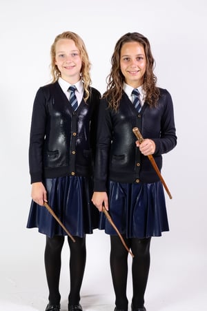 Two young wet soaked girls standing side by side against a white backdrop. Both are dressed in school uniforms, consisting of a dark-colored cardigan, a white shirt, and a navy-blue skirt and black wool tights. Completly soaked wet, wetness visible, They are also holding wands, reminiscent of those used in the Harry Potter series. The woman on the left has blonde hair, while the one on the right has wavy brown hair.