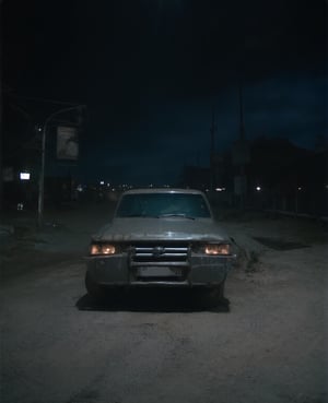 A truck parked on a deserted street at night, its headlights casting an eerie glow. The darkness of the surrounding buildings and the absence of any nearby streetlights emphasize the isolation of this late-night setting. The truck's metal body reflects the faint city lights, giving it a metallic sheen.