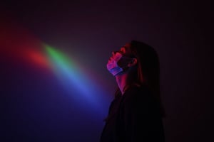 CridLight, cover face mask,
A moody, atmospheric portrait of a person, captured in a side profile. The subject is illuminated by a radiant beam of light that cuts across the scene, casting a spectrum of colors ranging from deep reds and purples to bright blues and yellows. The person's pose is contemplative, with their head tilted upwards and eyes closed, suggesting a moment of introspection or wonder. The individual is dressed in a dark, possibly black, outfit that contrasts with the luminous background. The lighting creates a dramatic and ethereal atmosphere, emphasizing the interplay of light and shadow. The background is dark, which accentuates the luminosity of the light and the subject, creating a sense of depth and dimension. The overall style of the image is evocative and evocative, with a focus on mood and atmosphere over detailed realism.