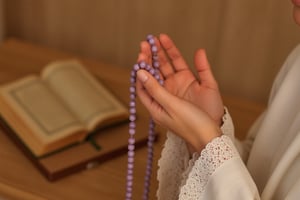 A full-body frontal shot of a woman crying heavily in prayer. Her hands are gently raised, cupped together with palms facing upward, conveying peace and devotion. She delicately holds a string of purple prayer beads in her right hand, with her fingers loosely aligned and slightly relaxed. She wears a white telekung, gracefully covering her wrists. The softly lit background features an out-of-focus Quran with a decorative cover, enhancing the spiritual atmosphere. The lighting is warm and natural, creating a serene and reverent ambiance,cinematic dramatic color style