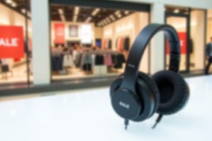 Sleek black headphones on a white background, slightly tilted to emphasize design, with a blurred department store in the background symbolizing a sale. Shot in stock photography style with shallow depth of field, natural lighting.