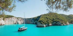 Captured from a high-angle perspective on a sunny day, a black sailboat is anchored in the middle of a body of water. The boat is adorned with a white sail, and a few people are seated on the deck. The water is a vibrant turquoise color, and the boat is floating on the surface of the water. To the left of the boat, a large tree branch is visible, adding a pop of color to the scene. In the background, a rocky cliff face is covered in lush green trees. The sky is a muted blue, with a few wispy clouds.,noc-landscape