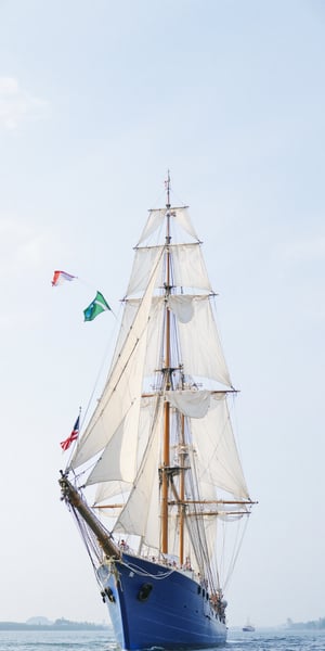 Captured at eye-level on a sunny day, a large sailing ship is seen in the middle of the frame. The ship is adorned with a blue body, adorned with white sails, and numerous people are seated on the bottom of the ship. The sails are adorned with two American flags, one red, white and blue, and the other green. The sky is a pale blue, dotted with wispy white clouds, adding a touch of color to the scene.