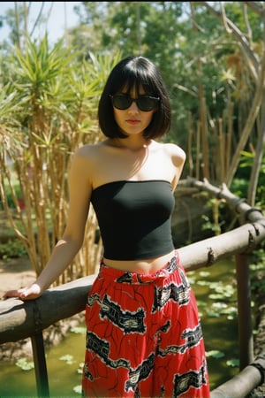 analog photo, a petite woman  with a bob haircut and bangs stands by a rustic fence in a lush, tropical outdoor setting, black hair, white skin, She wears a fitted black top and brightly colored boho-style pants with bold patterns in red, black, and white, Her oversized sunglasses and confident posture add to the modern and chic summer look, The background features palm trees, bamboo accents, and water plants, enhancing the tranquil and nature-filled atmosphere of the location, The sunlight and shadows give the scene a warm and inviting feeling, fade film, 100mm photo, grainy, vintage, stained, highly detailed, 100NewMemories