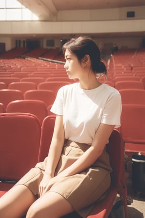 In a spacious, empty auditorium filled with rows of red and beige seats, a young woman sits alone, resting her head on her hand with a contemplative expression. Dressed casually in a light-colored t-shirt and skirt, she holds a can of soda, suggesting she might be waiting for an event to start or simply passing the time. The vastness of the empty seats around her emphasizes a sense of solitude and introspection, creating a poignant contrast between her quiet presence and the otherwise vacant, echoing space.