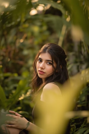 A stunning portrait of Kalyani Priyadarshan, framed by a warm golden light. She sits serenely in a lush greenery-filled setting, her long hair cascading down her back like a gentle waterfall. Her eyes sparkle with quiet confidence as she gazes softly into the distance.