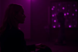 cinematic, A young woman with blonde hair sits in a dimly lit room, illuminated by soft purple and pink lighting. She gazes thoughtfully into the distance, with a bokeh of lights in the background, creating an intimate and contemplative atmosphere., film grain, Short telephoto focal length, shot on ALEXA 65