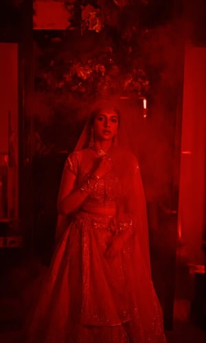 In a dimly lit, smoke-filled room, Madonna Sebastian poses majestically against a lavish, crimson-hued backdrop adorned with gleaming gold accents and subtle neon highlights. Her stunning red lehenga shimmers with intricate embroidery, paired with opulent gold jewelry that catches the eye. The cinematic lighting casts a warm, moody glow, evoking the essence of the iconic Matrix franchise. Madonna Sebastian exudes confidence and glamour, her regal presence commanding attention in this striking, neo-noir inspired setting.