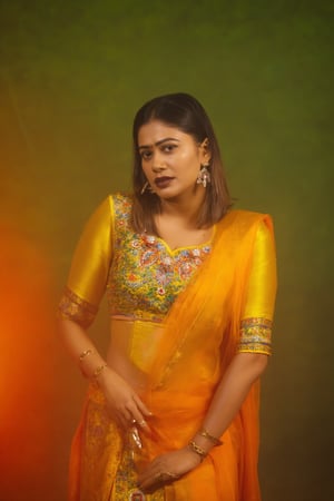 a woman dressed in a traditional Indian outfit stands in front of a vibrant orange backdrop. The woman's outfit is adorned with a yellow blouse, adorned with blue, red, and orange accents. She is wearing a pair of silver earrings, and a ring on her finger. Her hair is styled in a sleek bob, and her eyes are focused on the camera. She has a serious expression on her face, and she is wearing black lipstick. Her left hand is draped over her right shoulder, adding a touch of contrast to her outfit. The backdrop is a combination of green and orange, creating a striking contrast to the woman's body.,Payal