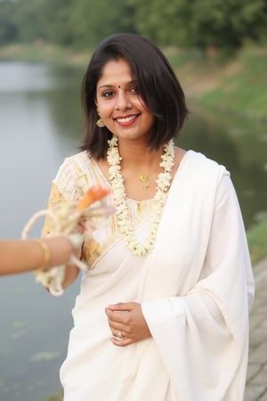 a woman dressed in a white saree adorned with gold bands and a gold necklace. She is adorned with a gold ring, earrings, and a garland of white flowers. Her hair is styled in a sleek bob, and she is smiling. The saree is draped over her left shoulder, adding a touch of contrast to her outfit. The backdrop is blurred, suggesting a natural setting.,Payal,Mallu beauty 