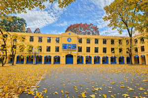 An outdoor setting, possibly a school or college campus, with a large yellow building in the background. The building has multiple arched windows and a blue emblem on its facade. In the foreground, there's a basketball court covered with fallen leaves, with a basketball hoop visible. The ground is mostly covered with dried leaves, and there are a few trees surrounding the court. The atmosphere seems calm and serene, with no people in sight.