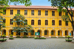 A vibrant yellow building with multiple windows, situated amidst a lush green courtyard. The courtyard is adorned with tall trees, their branches forming a canopy overhead. In the center of the courtyard, there's a paved area with a portrait of a man displayed prominently on the building's facade. The building has a classic architectural style with arched windows and decorative elements. The overall ambiance of the place is serene and inviting.