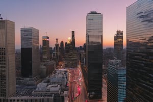 A sweeping aerial view of a metropolitan cityscape at sunset. Tall skyscrapers rise from the concrete jungle, their glass facades reflecting hues of orange and pink as the sun dips below the horizon. The streets are bustling with evening commuters, while neon lights and billboards illuminate the urban landscape.