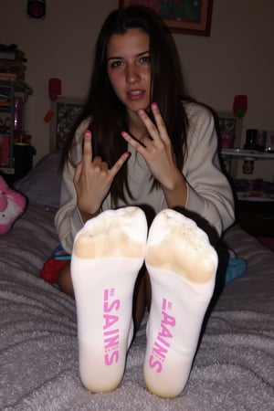 The image is a photograph of a young woman with a light skin tone and long, dark hair, posing in a cozy, cluttered bedroom. She is positioned on a plush, grey shaggy rug, with her feet prominently displayed in the foreground. She is wearing white socks with the word "VANS" printed in pink on the soles, which are worn and dirty, in fact the suggestion of her footprint appears in the dirt. suggesting she has been wearing them for some time. She is making the "L" sign with her fingers. 