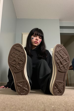 The image is a photograph of a young woman with a distinctive style, taken from a low-angle perspective, focusing on her feet and the soles of her converse all star shoes. She is sitting on a beige carpeted floor in a room with light grey walls and a textured, white ceiling. The woman has long, dark hair with bangs and is wearing a dark, casual outfit, including a hoodie and pants. She has stickers of cartoon characters plastered on her face. 