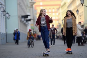 two young girls are riding skateboards on a cobblestone street. The girl on the left is wearing a red jacket with the word "Newport" written on the front, and a pair of blue jeans. Her hair is pulled back in a ponytail, and she is smiling. She is also wearing a gray t-shirt, a brown jacket, black pants, and orange sneakers. Her left foot is on a skateboard, and her right foot is in the air. The background is blurred, but it is a sunny day.