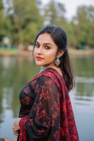 Captured at eye-level, a beautiful Indian woman, dressed in a black and red saree, stands in front of a body of water. Her hair is pulled back in a ponytail, and she's wearing a pair of silver earrings. Her left hand is resting on her hip, and her right hand is draped over her right shoulder. The backdrop is blurred, and the backdrop is a blend of green and brown trees.