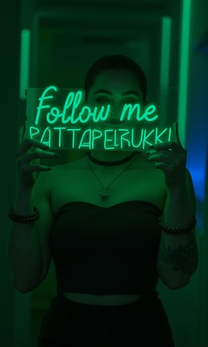 a vibrant young woman stands proudly holding a neon sign that reads "Follow me PATTAPERUKKI" in bold, glowing green letters. She is wearing a black strapless shirt, adorned with a black necklace and a black bracelet. Her left arm is adorned with tattoos, adding a pop of color to her outfit. The backdrop is a vibrant mix of green and blue lights, creating a striking contrast to her figure.