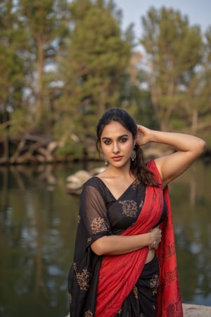 Captured at eye-level, a beautiful Indian woman, dressed in a black and red saree, stands in front of a body of water. Her hair is pulled back in a ponytail, and she's wearing a pair of silver earrings. Her left hand is resting on her hip, and her right hand is draped over her right shoulder. Exposed navel,  and the backdrop is a blend of green and brown trees.