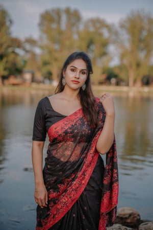 Captured at eye-level, a beautiful Indian woman, dressed in a black and red saree, stands in front of a body of water. Her hair is pulled back in a ponytail, and she's wearing a pair of silver earrings. Her left hand is resting on her hip, and her right hand is draped over her right shoulder. The backdrop is blurred, and the backdrop is a blend of green and brown trees.