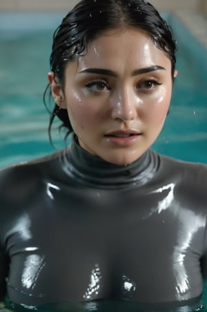 Daniella Pineda, emerging from the water. Her head and shoulders are visible above the surface of water. She looks surprised, her dark grey turtleneck top wet and clinging to her, with water droplets sparkling around her. She is in the middle of an indoor pool and splashes from her emergence. 