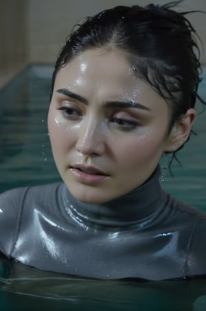 Daniella Pineda, emerging from the water. Her head and shoulders are visible above the surface of water. She looks scared and sad, her dark grey turtleneck top wet and clinging to her, with water droplets sparkling around her. She is in the middle of an indoor pool and splashes from her emergence. 